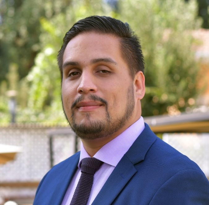 Hombre de pelo oscuro y corto con bigote y barba. Viste camisa, corbata y vestón.